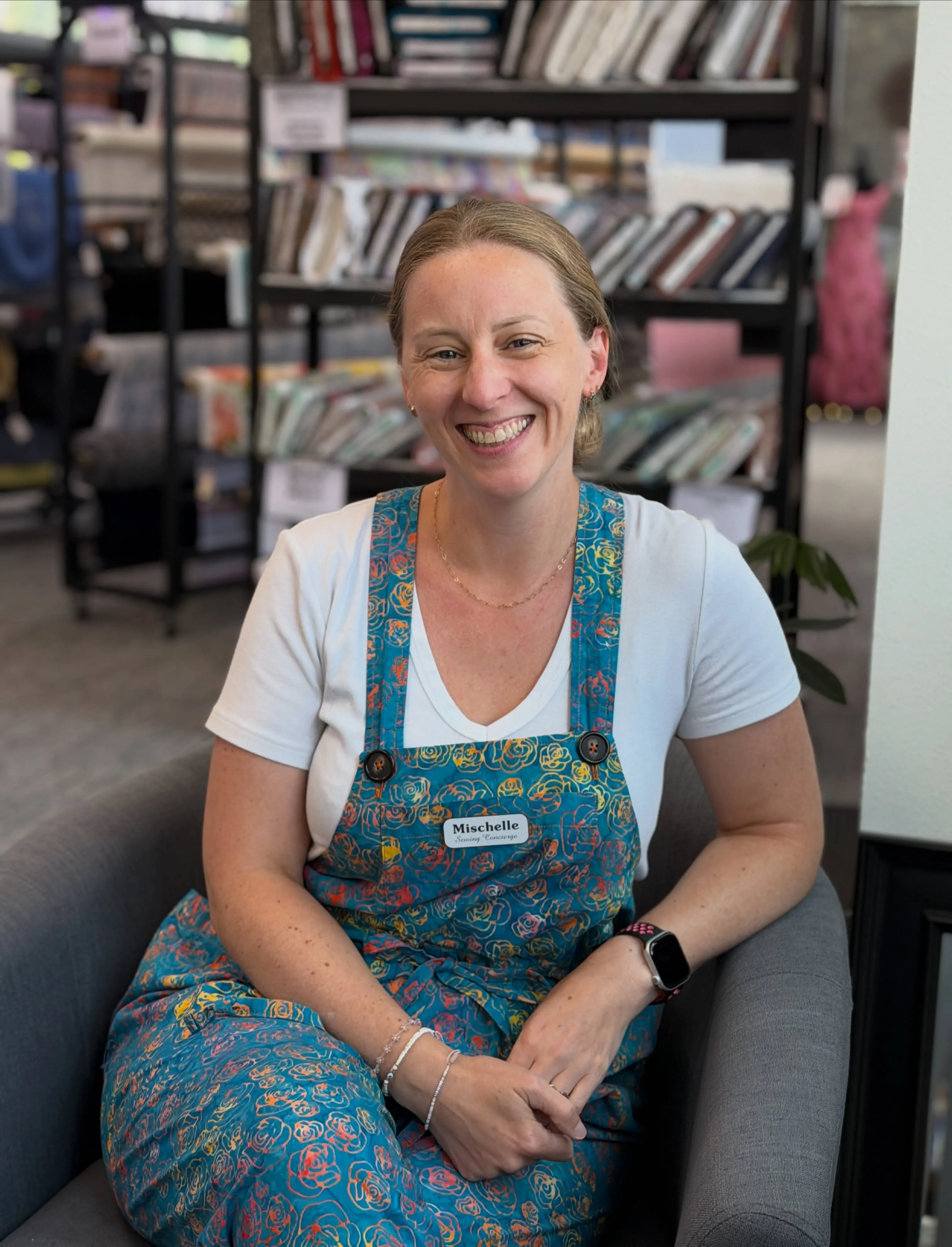 Mischelle Jillene, smiling, seated in her studio in front of shelves of fabric and pattern books.