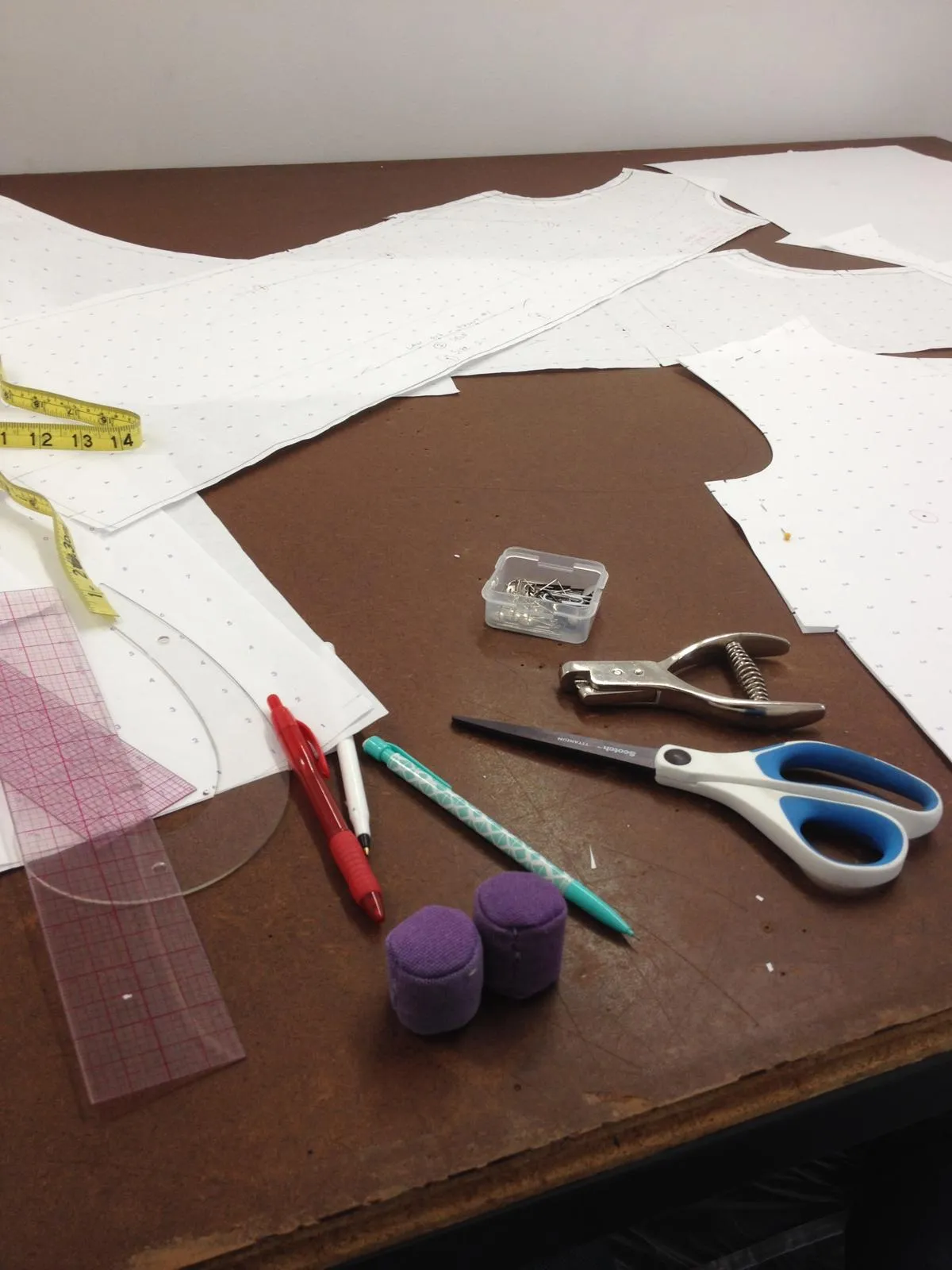 Pattern pieces drafted on dot-and-cross paper, with a tape measure, French curve, pins, and shears on the cutting table.